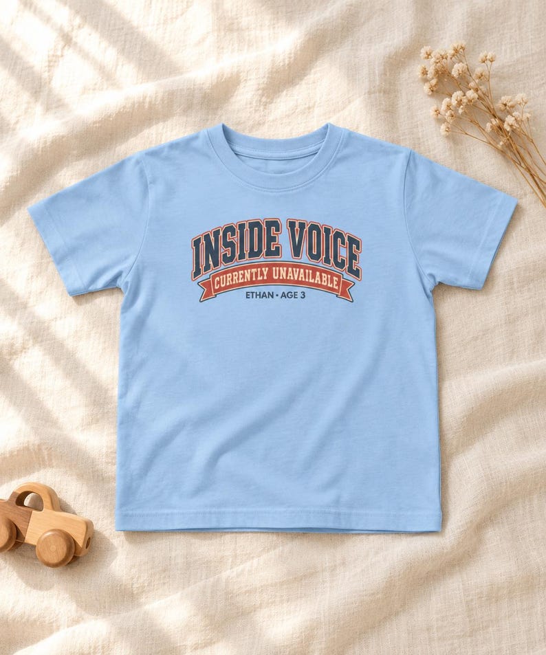 May include: A light blue short-sleeve t-shirt featuring a graphic with the text "INSIDE VOICE" in dark blue, "CURRENTLY UNAVAILABLE" in white on a red banner, and "ETHAN &bull; AGE 3" in dark blue lettering centered on the chest area.