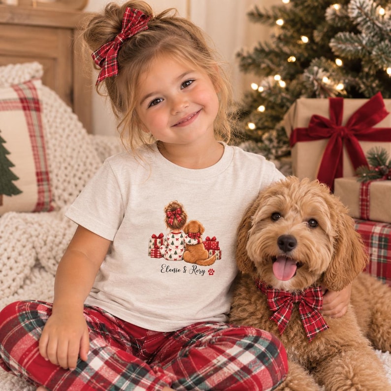 May include: A light heather gray short-sleeve t-shirt featuring a graphic of a child with a hair bow and a dog sitting by Christmas gifts. The text "Elonie & Rory" is printed below the graphic. The child wears a red plaid hair bow.