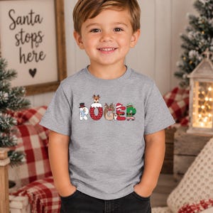 May include: A heather gray short-sleeve t-shirt featuring the name ROGER printed in large, festive letters. Each letter is decorated with Christmas-themed graphics, including a snowman, a reindeer, a Santa hat, and an elf outfit design.