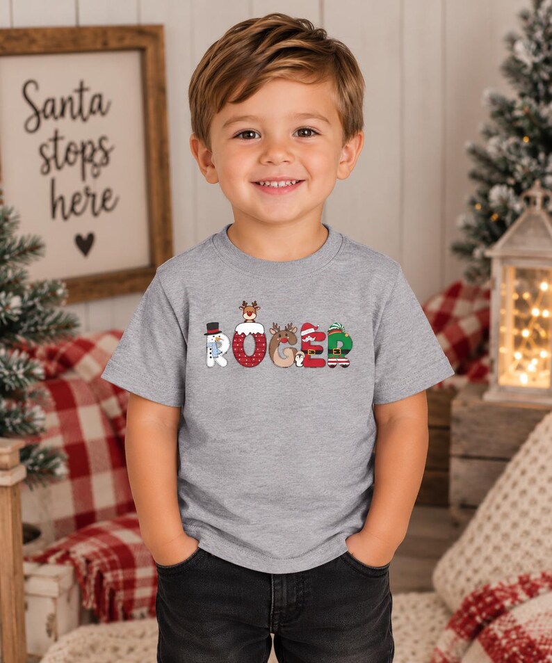 May include: A heather gray short-sleeve t-shirt featuring the name ROGER printed in large, festive letters. Each letter is decorated with Christmas-themed graphics, including a snowman, a reindeer, a Santa hat, and an elf outfit design.