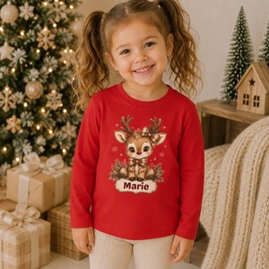 May include: A long-sleeved red shirt featuring a graphic illustration of a cartoon reindeer with a plaid bow and antlers, centered above a white name tag with the text Marie printed in brown, surrounded by small snowflakes and pine branches.