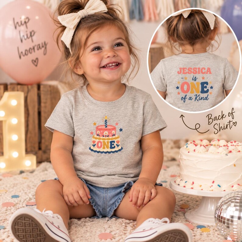 May include: A heather gray short-sleeve t-shirt featuring a graphic of a two-tiered birthday cake with the text ONE. The back of the shirt displays the text JESSICA is ONE of a Kind in colorful retro-style lettering with star accents.
