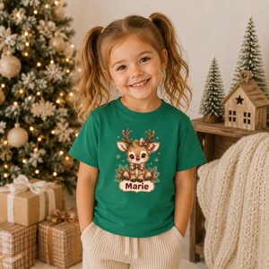 May include: A green short-sleeved t-shirt featuring a graphic of a cartoon reindeer with a plaid bow and scarf. The reindeer is surrounded by small snowflakes and pine branches. Below the reindeer, the name Marie is printed in brown text.