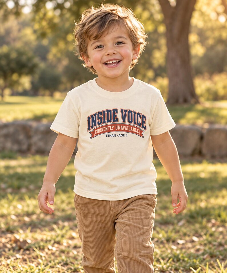 May include: A cream-colored short-sleeve t-shirt featuring the text INSIDE VOICE in dark blue with a red outline, followed by the text CURRENTLY UNAVAILABLE on a red banner, and the text ETHAN AGE 3 in dark blue lettering centered below.