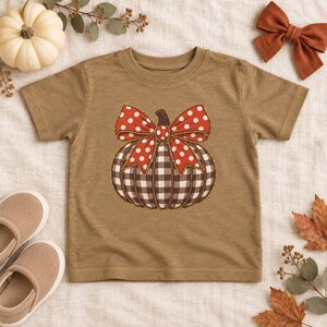 May include: A tan short-sleeve t-shirt featuring a central graphic of a pumpkin with a black and white buffalo plaid pattern, topped with a large red bow decorated with white polka dots, outlined in a thin gold stitch detail.