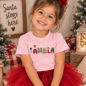 May include: A light pink short-sleeved t-shirt featuring the name PAMELA printed in a festive holiday-themed font. Each letter is decorated with Christmas motifs including a snowman, reindeer, Santa Claus, and elf-inspired patterns.