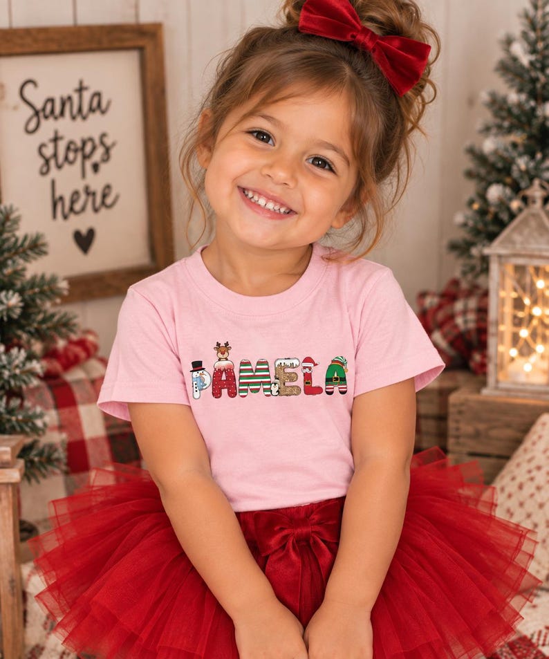 May include: A light pink short-sleeved t-shirt featuring the name PAMELA printed in a festive holiday-themed font. Each letter is decorated with Christmas motifs including a snowman, reindeer, Santa Claus, and elf-inspired patterns.
