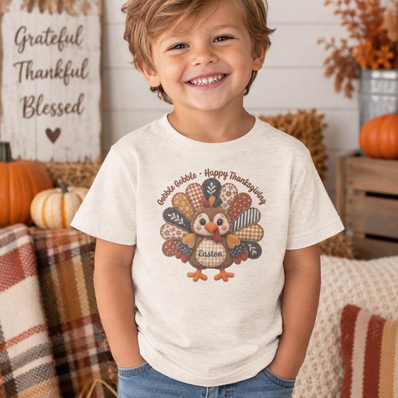 May include: A short-sleeved, heathered cream t-shirt featuring a central graphic of a turkey with colorful, patterned patchwork feathers. The text "Gobble Gobble - Happy Thanksgiving" arches above the turkey, and the name "Easton" is printed on its body.