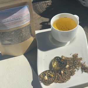 May include: A cup of herbal tea sits on a white plate with a heart-shaped tea infuser and loose tea leaves. A bag of "Tranquil Meadow" herbal tea is next to the plate. The tea is a light amber color.