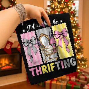 Peut inclure: T-shirt noir avec l'inscription "I'd rather be THRIFTING". Le motif comprend trois rectangles en forme de timbre-poste avec des nœuds et un chariot de magasinage rempli d'objets vintage. En arrière-plan, une cheminée et un sapin de Noël.