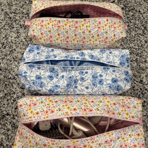 May include: Three rectangular cosmetic bags with zippers. Two bags have a white floral pattern with pink and yellow flowers. The third bag has a blue floral pattern. The bags are open, revealing hair styling tools inside.