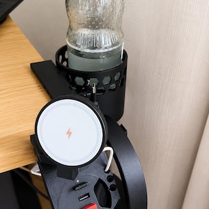 Pode incluir: Um suporte de mesa preto com uma base de carregamento sem fio circular com ícone de raio, um porta-copos com padrão de favo de mel, um gancho para fones de ouvido e um hub com portas USB e um botão vermelho, marcado com o texto Hama.