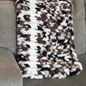 May include: A handmade, chunky knit blanket in a mix of white, brown, and black. The blanket is folded over the arm of a gray sofa. The thick yarn creates a textured, cozy appearance, perfect for home decor.