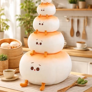 Puede incluir: Una pila de cuatro peluches redondos, blancos y naranjas que se asemejan a bollos al vapor. Cada uno tiene una cara con ojos pequeños y una boca, y pies naranjas. Los juguetes están sobre una mesa de madera con un mantel individual de color neutro.