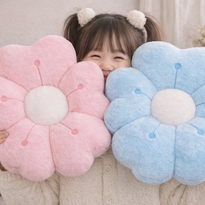 May include: Two flower-shaped cushions, one pink and one blue, with white centres and embroidered details. The cushions are soft and fluffy, ideal for home decor or as a gift.