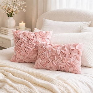 Può includere: Due cuscini decorativi rosa con un design a petali poggiano su un letto bianco. I cuscini sono quadrati e rettangolari e i petali creano una superficie strutturata. Sono visibili anche una coperta bianca e altri cuscini bianchi.