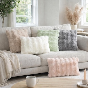 May include: A selection of faux fur throw cushions in shades of beige, white, pale green, grey, and pink. The cushions are arranged on a light grey sofa. A beige throw and a coffee cup are also visible. The cushions are designed for home decor.