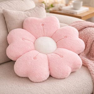May include: A soft, pink flower-shaped cushion with a white central disc. The cushion has six petals and is made of a fluffy material. It rests on a light-coloured sofa, with other cushions and a blanket visible in the background.