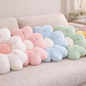 May include: A selection of flower-shaped cushions in pastel shades of white, pink, blue, green, and yellow. The cushions are arranged on a light-coloured sofa, creating a soft and inviting aesthetic.