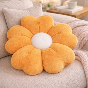 May include: A flower-shaped cushion with a white central disc and orange petals. The petals are heart-shaped and have a soft, textured appearance. The cushion is placed on a neutral-toned sofa.