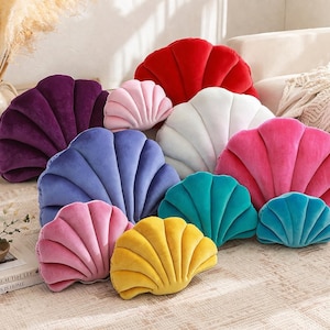 Puede incluir: Una colección de cojines con forma de concha marina en varios colores, incluyendo rojo, morado, rosa, azul, amarillo, blanco y turquesa. Los cojines están hechos de un material suave y aterciopelado y están colocados sobre una superficie estampada.