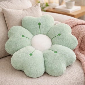 May include: A flower-shaped cushion with five light green petals and a white centre. Each petal has a small green accent. The cushion is on a beige sofa, with a pink blanket visible in the background.