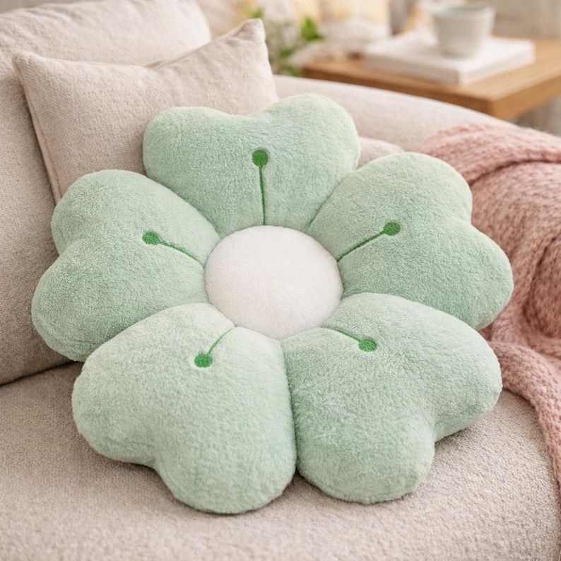 May include: A flower-shaped cushion with five light green petals and a white centre. Each petal has a small green accent. The cushion is on a beige sofa, with a pink blanket visible in the background.