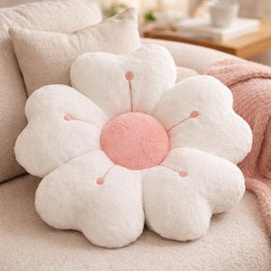 May include: A flower-shaped cushion with white, heart-shaped petals and a pink centre. The petals have small pink details. The cushion is placed on a beige sofa, creating a soft and inviting aesthetic. The cushion is a decorative item.