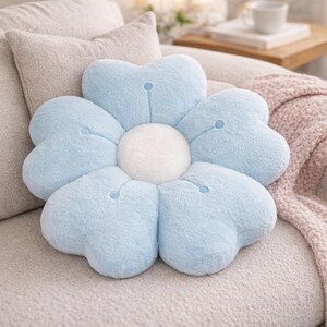May include: A light blue flower-shaped plush cushion with a white centre. The cushion has six heart-shaped petals. The cushion is on a light-coloured sofa with other cushions and a blanket in the background.