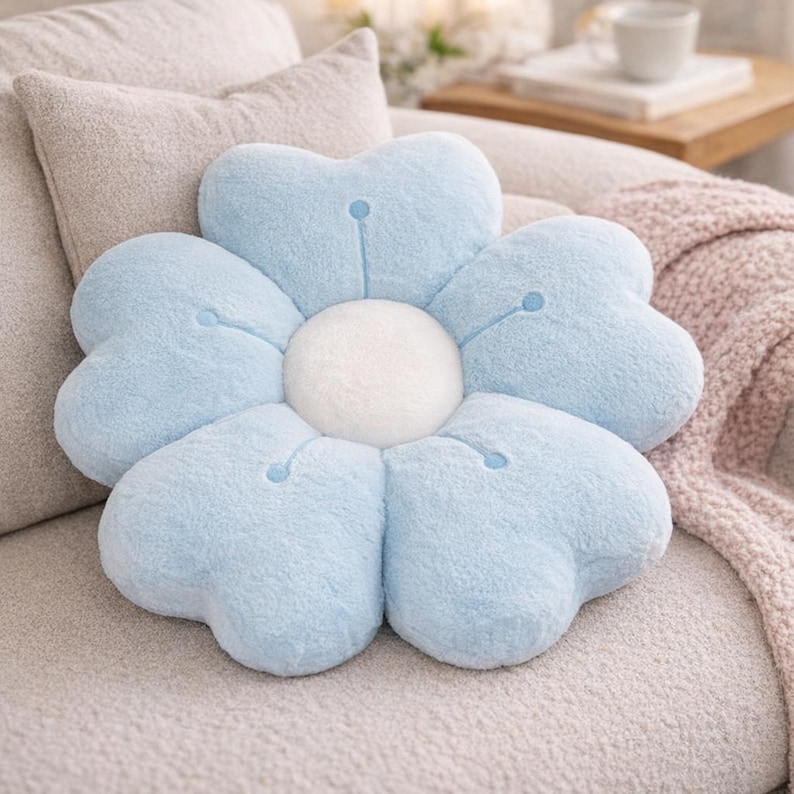 May include: A light blue flower-shaped plush cushion with a white centre. The cushion has six heart-shaped petals. The cushion is on a light-coloured sofa with other cushions and a blanket in the background.