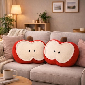 Puede incluir: Dos cojines con forma de manzana, rojos por fuera y blanco roto por dentro, cada uno con dos semillas negras y un tallo marrón. Están colocados sobre un sofá gris, creando un ambiente acogedor. Miden unos 40 cm de largo.