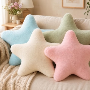 Può includere: Quattro cuscini a forma di stella in colori pastello: azzurro, crema, verde e rosa. I cuscini sono disposti su un divano beige, creando un'estetica morbida e accogliente. Questi cuscini decorativi aggiungono un tocco di fantasia.