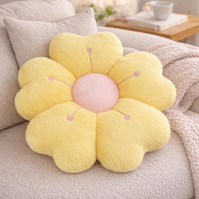 May include: A flower-shaped cushion with yellow petals and a pink centre. The cushion is on a light grey sofa. The petals have pink embroidered details. A decorative home accent.