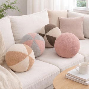 Pode incluir: Uma coleção de almofadas redondas decorativas em várias cores, incluindo rosa, castanho e creme, dispostas num sofá branco. As almofadas têm um aspeto texturizado e macio e são projetadas em forma esférica segmentada. Uma mesa de centro com uma caneca e livros está em primeiro plano.