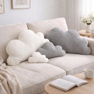 Könnte beinhalten: Eine Sammlung von wolkenförmigen Zierkissen in verschiedenen Größen und Weiß- und Grautönen. Die Kissen sind auf einem beigefarbenen Sofa angeordnet und schaffen eine gemütliche und skurrile Ästhetik. Der weiche, strukturierte Stoff sorgt für visuelles Interesse und Komfort.