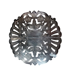 May include: An ornate silver-coloured metal decorative plate with a detailed floral and leaf design. The plate is circular with a central square area and a scalloped edge. The design includes flowers, leaves, and intricate scrollwork.