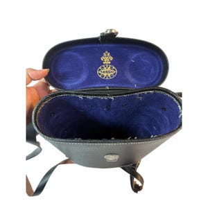 Pode incluir: Estojo Tasco em couro preto, aberto para revelar um interior de veludo azul. O estojo tem um fecho prateado e uma borda costurada. O interior apresenta o logotipo Tasco em dourado. O estojo tem uma alça preta.