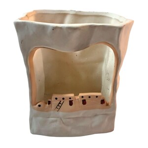 May include: A cream-colored ceramic planter with a rectangular opening and a cutout revealing a Southwestern-style village scene. The village features small, tan buildings with red accents and a ladder.