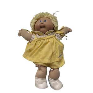 Puede incluir: Muñeca Cabbage Patch Kid vintage con cabello rubio de hilo y ojos azules. La muñeca viste un vestido de cuadros amarillos y bragas a juego. Zapatos blancos completan el atuendo. Los brazos de la muñeca están extendidos.