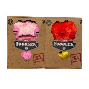 Op de afbeelding: Twee ZURU Fuggler pluche speelgoed in hun originele verpakking. Eén is roze en de andere is rood. De dozen zijn bruin met de woorden "LOVE FUGG" en een hartvormig pluche speelgoed.