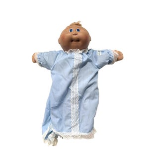 May include: A vintage Cabbage Patch Kids doll with a tan complexion, blue eyes, and light brown hair. The doll is wearing a light blue baby doll dress with white lace trim. The doll's arms are outstretched.