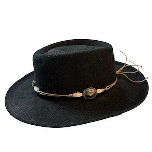 May include: Black fedora-style hat with a decorative band featuring a silver-toned oval medallion and off-white beads. The hat has a wide brim and a brown leather cord detail.