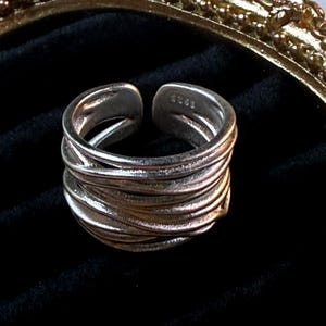 May include: Silver-toned adjustable ring with a layered, woven design. The ring features an open design and is set against a black background. The ring's texture and design create a unique, modern look. The ring is a piece of jewelry.