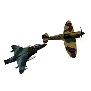 May include: Two toy airplanes on a white background. One is a green and gray Harrier jet with the word "MARINES" on the side. The other is a camouflage-patterned Spitfire with red, white, and blue roundels.