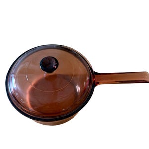 Puede incluir: Un cazo redondo de cristal color ámbar con una tapa de cristal transparente a juego. La tapa presenta un pequeño pomo circular de color marrón oscuro en el centro. El cazo tiene un mango largo de cristal integrado en un lado del borde.