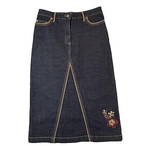 Puede incluir: Falda vaquera azul oscuro con abertura frontal y bordado floral decorativo cerca del dobladillo. La falda presenta costuras color canela y bolsillos con ribete color canela. Cierre de botón y cremallera en la cintura.