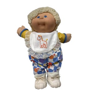 May include: A vintage doll with blonde, curly hair and blue eyes. The doll wears a bib with a horse design, a yellow and blue sleeved shirt, and blue, white, and purple checkered trousers. White trainers complete the outfit.