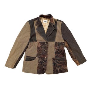 May include: A patchwork blazer featuring alternating panels of solid tan and dark charcoal denim fabric. The garment includes a floral pattern on specific sections, a notched lapel, three front buttons, and two lower patch pockets with a HUCKE label.
