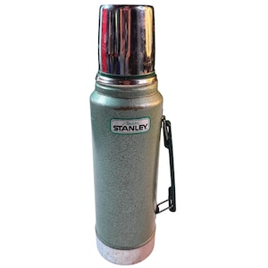 May include: A vintage green Stanley Aladdin thermos featuring a textured hammer-tone finish, a polished chrome cup lid, and a dark green plastic side handle. The front displays a white rectangular label with the text Aladdin STANLEY.