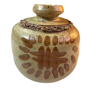Può includere: Un vaso in ceramica fatto a mano con coperchio. Il vaso è di colore beige chiaro con disegni floreali marroni e un bordo strutturato. Il vaso ha un corpo arrotondato e un piccolo collo cilindrico.